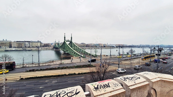 Obraz View of the road and tramway lines of Pest with the Liberty Bridge seen from the Gellert Hill rock chapel at a winter sunset. Buda cityscape on the other side of the Danube River, Budapest, Hungary.