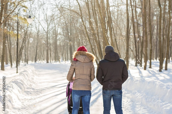 Fototapeta Happy young family walking in the park in winter. The parents carry the baby in a stroller through the snow.