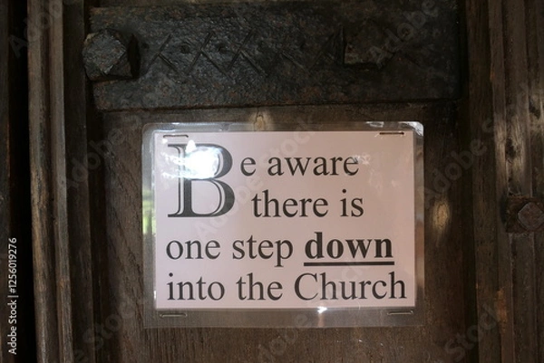 Fototapeta A small, weathered wooden sign stands at the church entrance, its black lettering clear against the aged surface:  Be Aware: One Step Down. The simple message, framed by a decorative border.