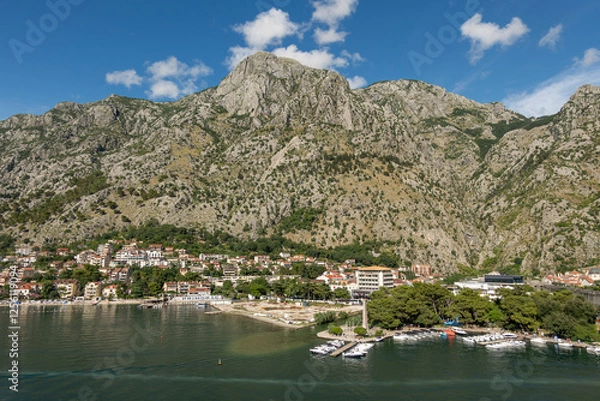 Fototapeta Kotor, Montenegro - July 2, 2024: Landscape, new and historic town with its ramparts and moat in front of tall mountain range under blue cloudscape