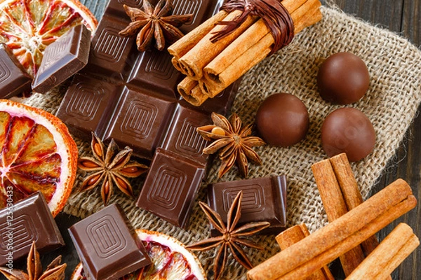 Fototapeta Dark and milk chocolates with dry oranges, candies, cinnamon and anise on burlap on rustic wooden table
