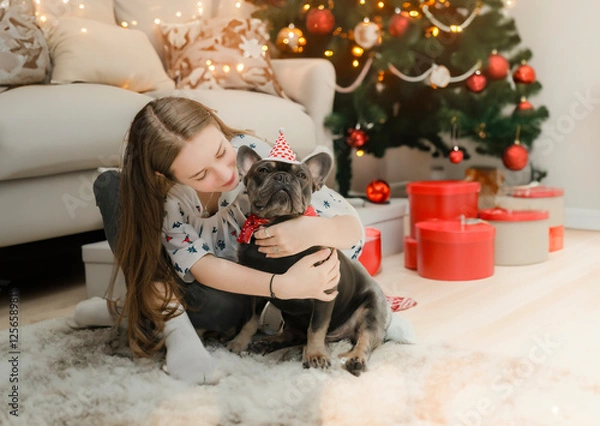 Fototapeta In a joyful and loving moment, a young girl and her dog smile together against a background of Christmas decorations.