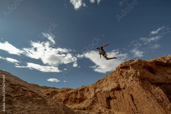 Fototapeta A man with trekking poles in hiking clothes jumps from one edge of a cliff to the other. A traveler's leap against the backdrop of a canyon and blue sky. Jump against the sky, view from the bottom.