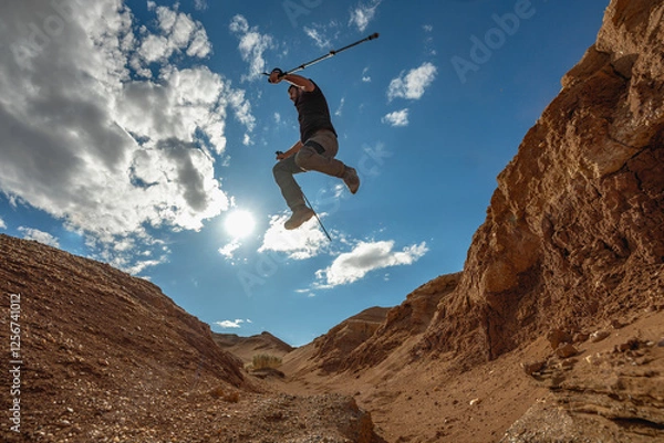 Fototapeta A man with trekking poles in hiking clothes jumps from one edge of a cliff to the other. A traveler's leap against the backdrop of a canyon and blue sky. Jump against the sky, view from the bottom.