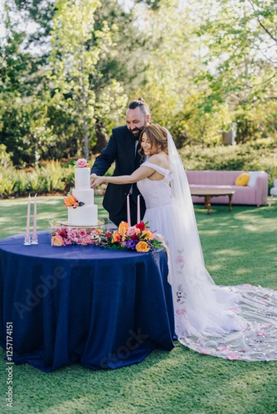 Fototapeta A joyful couple celebrates their love during a wedding cake cutting ceremony in a beautifully decorated garden, surrounded by loved ones and vibrant flowers, creating unforgettable memories for all