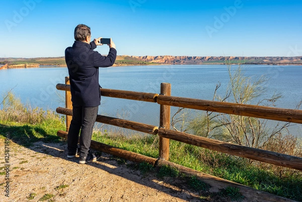 Fototapeta Man taking pictures with mobile phone of the landscape formed by the mountains and Lake Castrejon, Toledo, Spain.