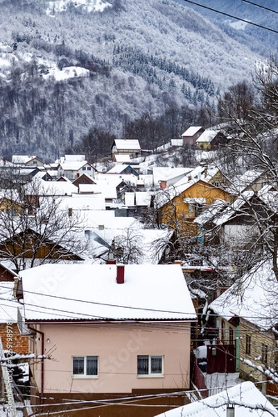 Obraz Different houses in snowy mountains during winter, surrounded by snow-covered trees. Pink house with snow covered roof. A peaceful scene. Vertical photo