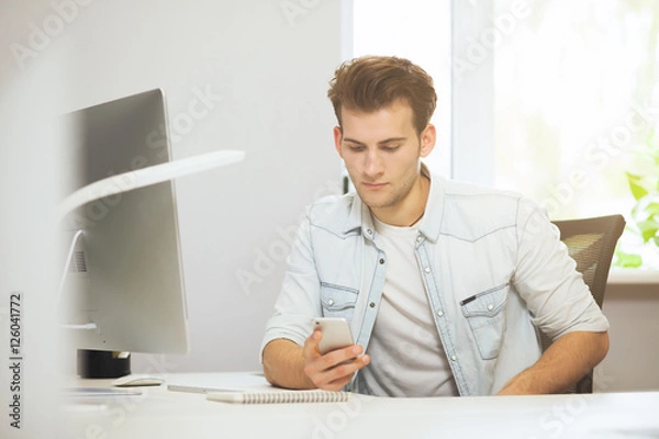 Fototapeta Portrait of handsome young businessman sitting at office table with personal computer and writing on mobile phone. Communication concept