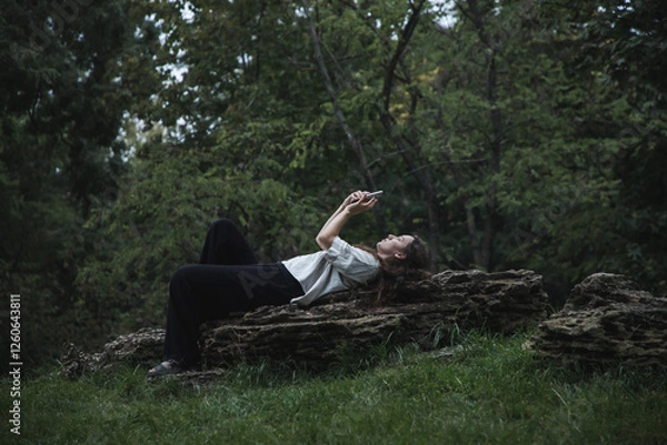 Obraz A girl is glued to her phone in nature. A girl can't relax without her phone. A girl reads the news, sits on social networks.