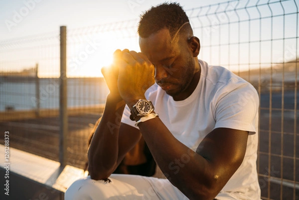 Obraz Young african american man praying with hands together at golden hour near fence