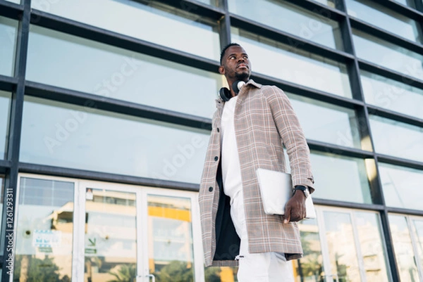Obraz Stylish entrepreneur walking with laptop in hand near modern office building