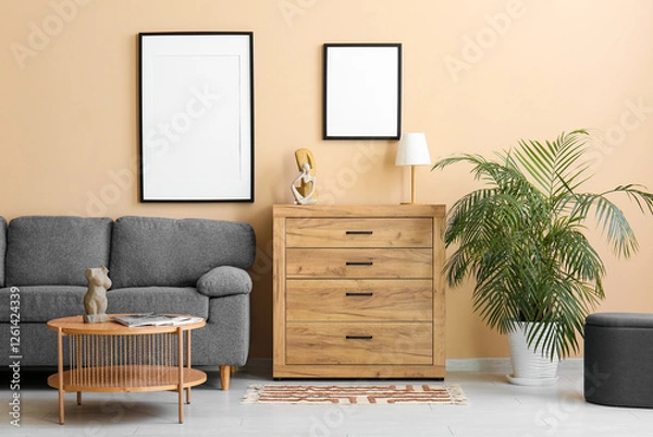 Fototapeta Chest of drawers, table and sofa near beige wall with hanging empty posters