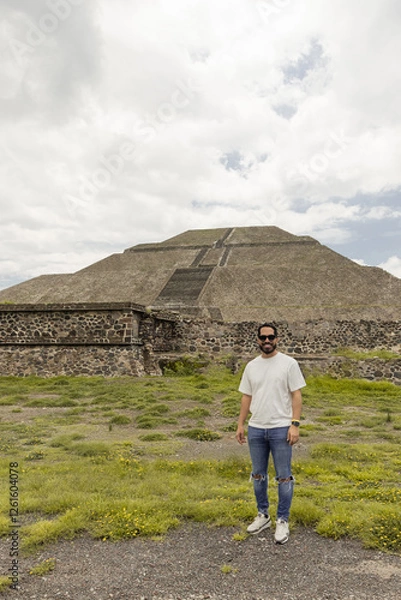 Fototapeta Teotihuacan, Mexico. A Mexican American tourist with a backpack and hat enjoys the view from the top of the Moon Pyramid. The Sun Pyramid and Avenue of the Dead can be seen in the distance.