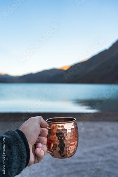 Fototapeta A solitary figure stands by the serene waters of El Yeso, Andes, Chile The hand, weathered and aged, clasps a copper mug, reflecting the pale light of the dawn in the mountains