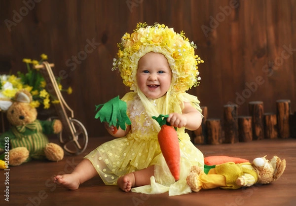 Obraz Portrait of a little girl in a yellow suit. Girl 7 months old sitting. Little child in a hat made of flowers. Little child holding a carrot in his hands