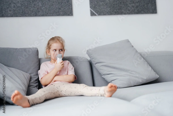 Fototapeta Portrait of a blond child doing inhalation on a gray sofa in the interior of the room. Lung health in children.