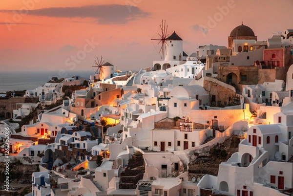 Fototapeta Santorini glows at sunset, with whitewashed houses cascading down rugged cliffs, windmills standing tall, and golden lights reflecting off the Aegean Sea, creating a dreamy Mediterranean scene.