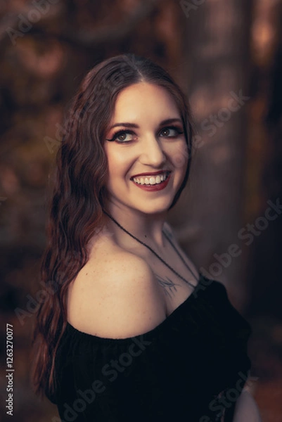 Fototapeta This striking photograph features a woman standing in a dark, moody forest, exuding a gothic and mysterious aura. She is dressed in an all-black ensemble, including a sheer, flowing cape.