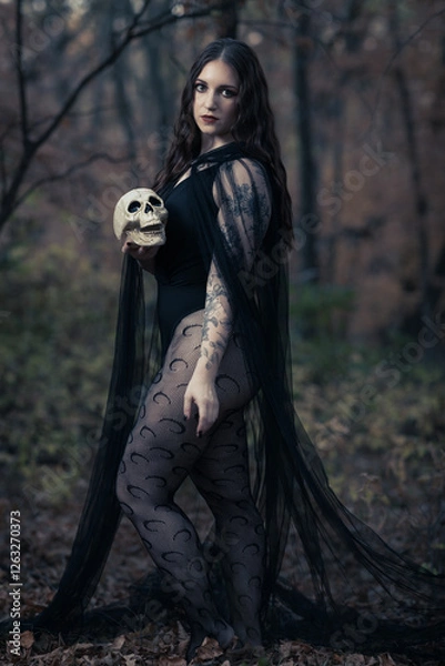 Fototapeta This striking photograph features a woman standing in a dark, moody forest, exuding a gothic and mysterious aura. She is dressed in an all-black ensemble, including a sheer, flowing cape.