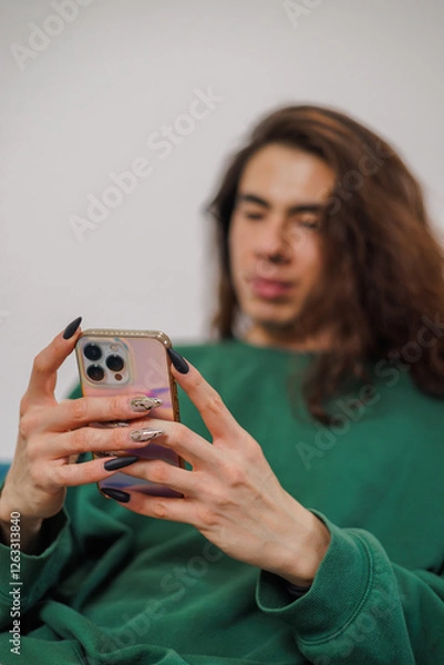 Fototapeta Non-binary person using their cell phone at home with blurred background. Long nails.