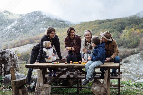 Fototapeta Cheerful friends with kids near the table against mountains in a