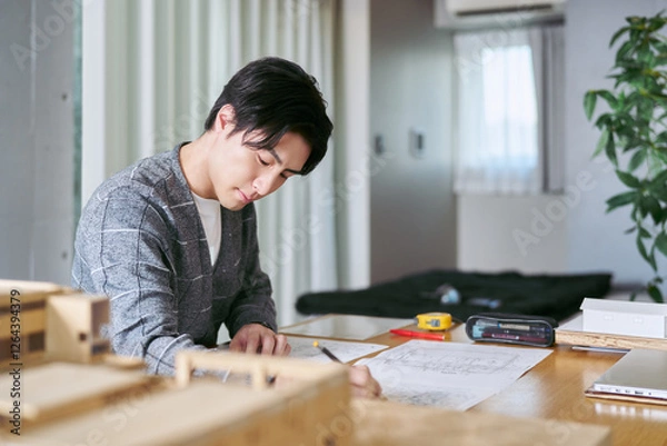 Obraz 自宅兼事務所で建築模型の置かれたテーブルで印刷された図面のチェックと修正をする建築士の男性