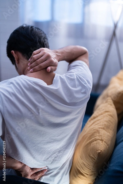 Obraz Asian freelancer man sitting on a couch holding his neck and lower back in discomfort, possibly due to muscle strain, poor posture, or stress and tired of working