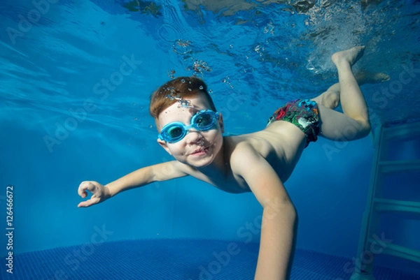 Obraz Little boy swims underwater in the pool, smiling, blowing bubbles and looking at me. The view from under the water. Close-up. Horizontal orientation