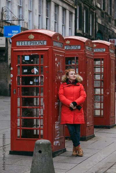 Fototapeta Tourist wearing a red coat exploring historic telephone booths in Scotland on a chilly day