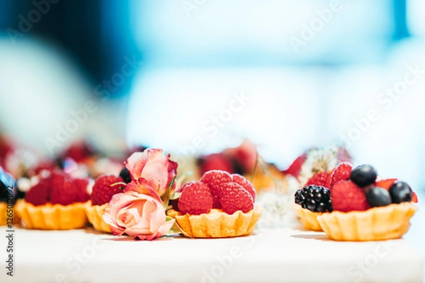 Fototapeta Nahaufnahme von kleinen Obsttörtchen, Tartelettes mit Beeren. Himbeeren, Blaubeeren, Brombeeren und essbaren Blumen, Rosen, Schleierkraut. Törtchen auf heller Oberfläche. Hintergrund unscharf