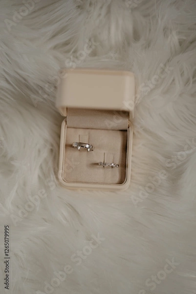 Obraz An elegant beige velvet ring box with a pair of wedding bands, including a sleek silver men's band and a delicate diamond-studded women's ring, placed on a soft white faux fur background