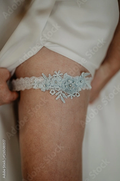Obraz  A close-up of a bride lifting her white wedding dress to reveal a delicate lace bridal garter adorned with intricate blue floral embroidery and sparkling rhinestones, symbolizing a timeless wedding 