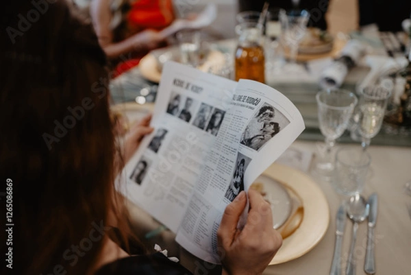 Obraz A guest at an elegantly set dining table attentively reads a black-and-white printed wedding newspaper featuring vintage photographs and articles, surrounded by fine glassware, gold-rimmed plates