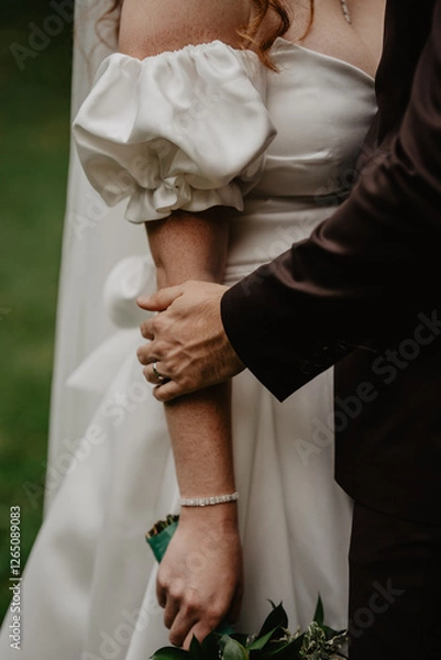 Obraz  A groom in a dark suit tenderly holding the bride’s arm, showcasing their wedding rings, as she wears a white satin dress with elegant puff sleeves, a delicate diamond bracelet, and holds a bouquet 