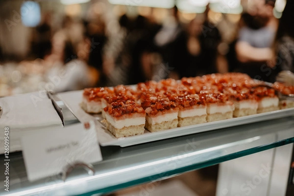 Obraz A tray of beautifully arranged layered dessert bars topped with fresh diced strawberries and glaze displayed on a glass counter at a catering event with blurred guests in the background