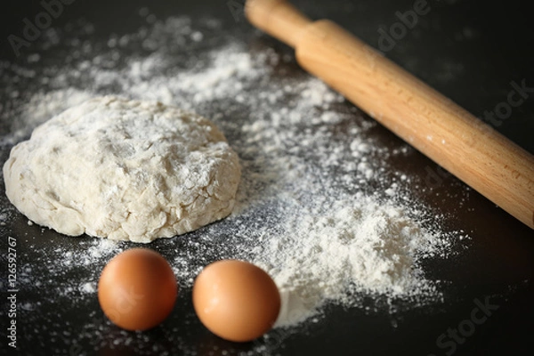 Fototapeta Dough with flour, eggs and rolling pin on dark table