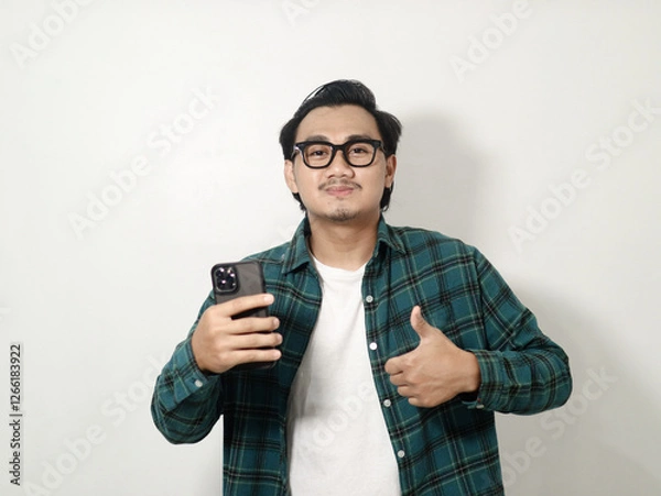 Obraz Smiling satisfied young Asian man wearing a plaid checkered shirt holding a mobile phone and showing thumbs up sign, recommends apps isolated on white background