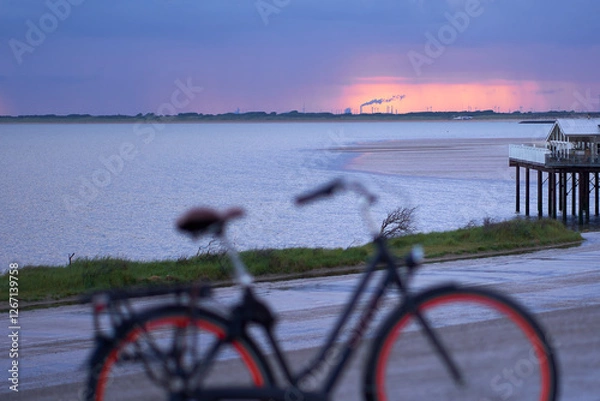 Fototapeta Port building of Rotterdam on horizon at sunrise. Stilt house at the water's edge of the North Sea. Ladies bicycle in depth blur. Netherlands, Zeeland, Brouwersdam.