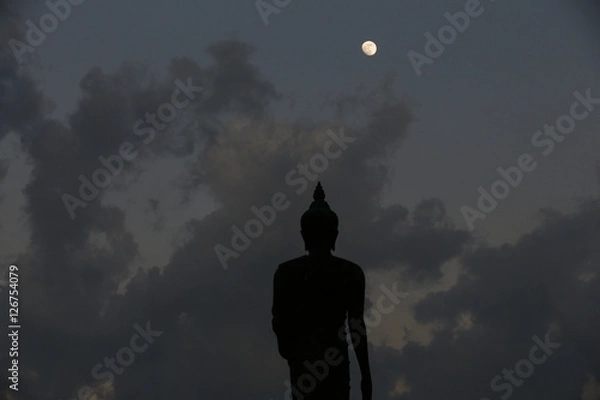 Fototapeta A moon shines in a sky over a large silhouetted Buddha statue in Bangkok, Thailand.