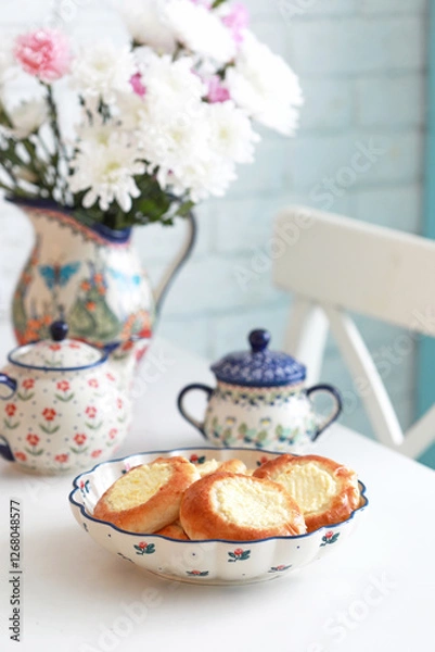 Obraz Ceramic dish with cheesecakes on a white kitchen table. There is a jug of flowers and a teapot in the background. Selective focus.