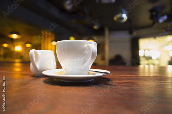 Fototapeta White cup of coffee on a saucer on a table in a dark cafe