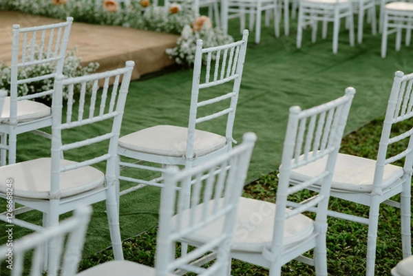 Fototapeta White chairs placed in a wedding setting in the garden and grass. Brown stage decorated with flowers