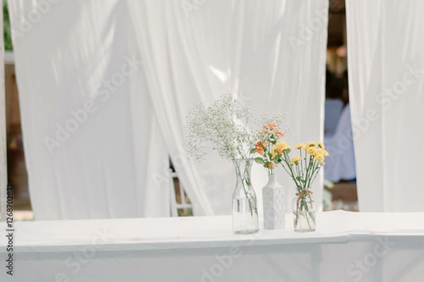 Fototapeta Fresh flowers in three vases are used for wedding decorations placed on a table covered with a white tablecloth. Additionally, there is a white cloth adorning the backdrop