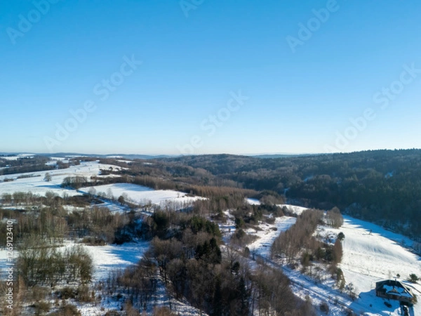 Fototapeta Aerial view of snow-covered fields and trees under a clear blue sky, showcasing a serene winter landscape. Perfect for nature-related projects.