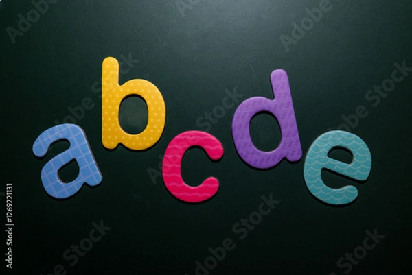 Fototapeta Colorful foam letters “a,” “b,” “c,” “d,” and “e” arranged on a dark background, symbolizing early learning, literacy, and creative design. Perfect for educational or childcare materials.