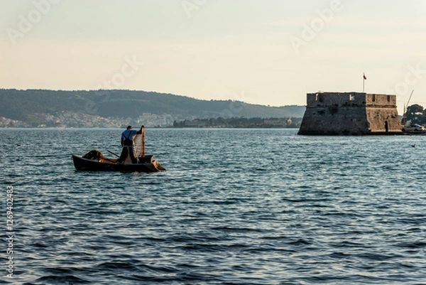 Obraz Fisherman on the Adriatic Sea in Croatia. A lone fisherman in a small boat pulls in his net at sunrise, with an ancient stone fortress and distant hills creating a peaceful coastal scene.