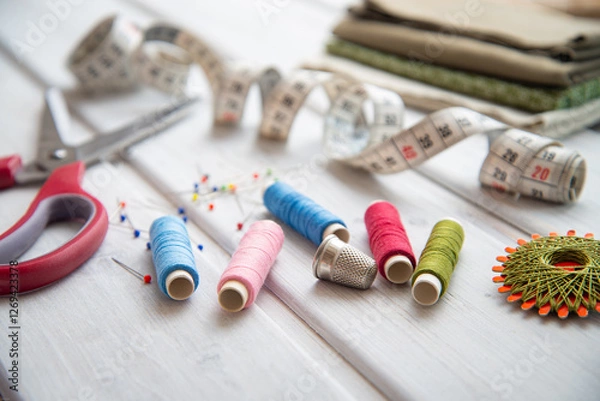 Fototapeta Sewing utensils - colored spools of thread, fabrics, tape measure and thimble on white wood