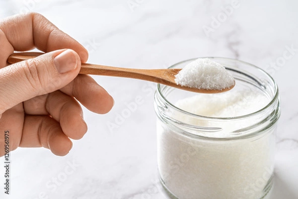 Fototapeta Woman fingers hold wooden spoon full of monosodium glutamate white crystals over glass jar. Favor enhancer MSG, food additive E621 concept.