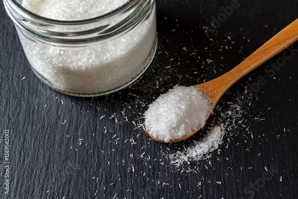 Obraz Monosodium glutamate in a wooden spoon and glass jar full of MSG white crystals over black background. Concept of food additive flavor enhancer E621.