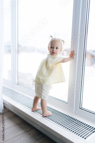 Fototapeta Vertical shot of adorable toddler girl with two playful ponytails standing by window on windowsill, soaking in winter scenery enjoying cozy warmth of home. Charming scene of childhood innocence.